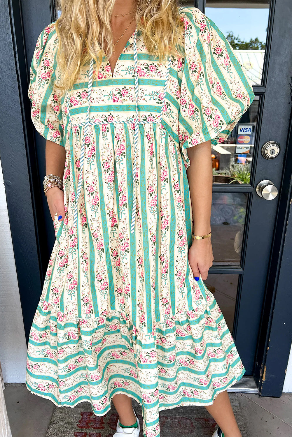 A close-up shot of a bohemian-style floral maxi dress with a tiered skirt. The dress displays a mix of earthy tones like mustard and olive, with delicate pink and purple floral patterns scattered across the fabric